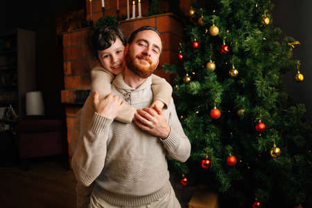 Medium shot portrait of happy Caucasian father and child son hugging on background of Christmas tree...