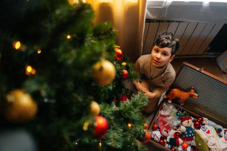 High-angle view of adorable child boy decorating Christmas tree with shiny balls and toys, sitting...