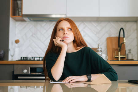 Front view of confused young woman writing handwritten letter sitting at table with envelope in...