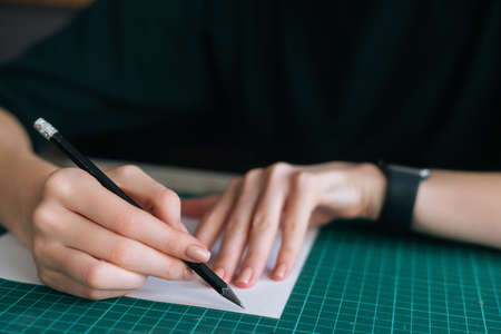 Close-up hands of unrecognizable female designer making mark with pencil on white blank paper lying on green rubber cutting mat at desk, selective focus. Concept of creative work and hobby.の写真素材