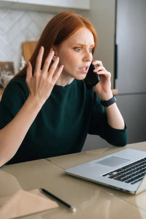 Vertical shot of furious young woman talking on mobile phone and using laptop sitting at table in kitchen with modern interior. Annoyed female freelancer talking on cellphone with manager.の写真素材