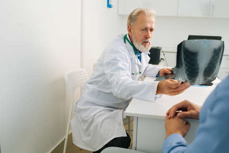 View from back of unrecognizable male patient listening mature adult doctor with chest x-ray lungs scan giving professional medical consultation for treatment illness, explaining causes of disease.の写真素材