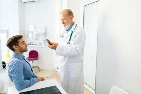Standing mature male adult doctor with smartphone consulting sick male patient sitting at table in medical office. Handsome young man client listening to older senior physician explaining treatment.の写真素材
