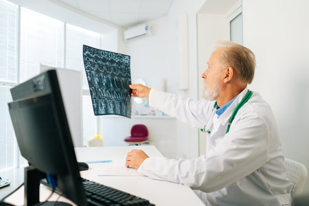 Side view of focused senior adult male doctor analyzing MRI spine image of patient sitting at desk...
