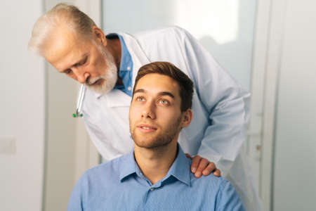 Close-up of mature adult male doctor chiropractor osteopath talking with young man patient sitting in chair, asks about problems. Senior manual therapist consulting patient about illness.の写真素材