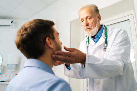 Close-up of mature adult male doctor otolaryngologist touching lymph nodes on neck and examining throat of male patient during medical checkup. Sick young man on consultation with sore throat.の写真素材