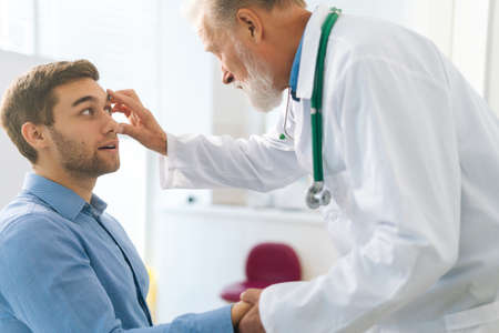 Close-up of mature adult male ophthalmologist examining eyes of male patient during checkup visit in clinic office. Young man coming in for consultation before undergoing vision correction surgery.の写真素材