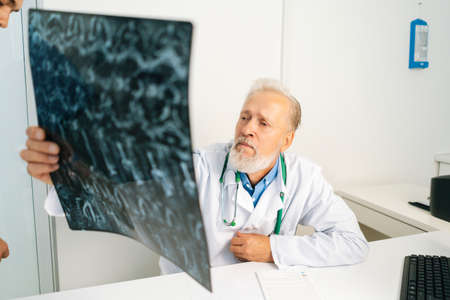 Pensive mature adult male doctor looking MRI spine image of patient sitting at desk with computer in...