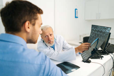 View from back of young male patient listening serious mature adult doctor explaining results of MRI scanning, pointing on image, giving professional consultation for treatment illness,の写真素材