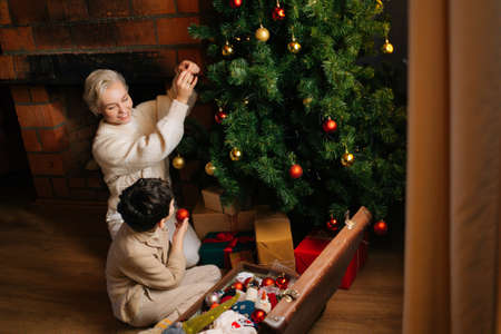 High-angle view of smiling young mother and child son decorating with colorful balls of Christmas tree sitting on floor near fireplace. Happy family preparing house for New Year holidays celebration.の写真素材