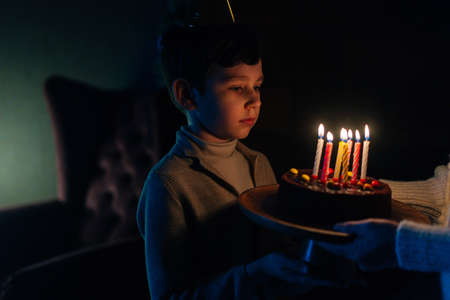 Close-up of adorable little child boy making wish before blowing candles on birthday celebration cake in dark room. Cheerful Caucasian boy looking on burning candles on cake at home.の写真素材
