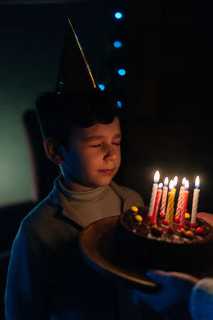 Vertical close-up shot of happy child boy with closed eyes making wish before blowing candles on birthday celebration cake in dark room. Unrecognizable mother holding cake for beloved kid at home.の写真素材