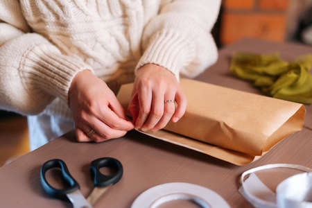 Close-up of unrecognizable young woman wrapping festive Christmas gift box in craft paper with presents for family and friends. Preparing for xmas, new year and winter holidays, festive moodの写真素材