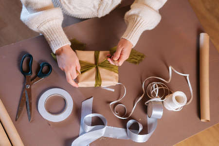 Close-up high-angle view of unrecognizable young woman tying ribbon on wrapped Christmas gift box with presents for family and friends. Preparing for xmas, new year and winter holidays, festive mood.の写真素材