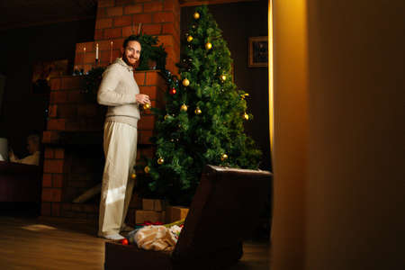 Cheerful handsome young man in cozy sweater decorating Christmas tree near fireplace in dark living room, looking at camera. Caucasian bearded male preparing home for New Year holidays celebrationの写真素材