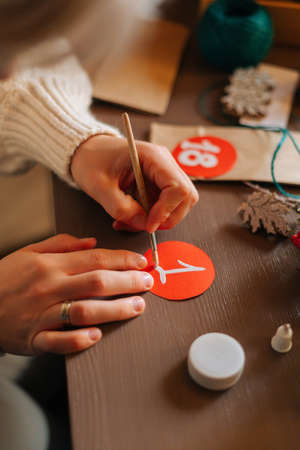 High-angle view of vertical shot of young woman writing number on red bag by brush and white paint sitting at table. Happy female making paper bags from kraft paper for advent calendar on xmas Eve.の写真素材