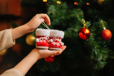 Close-up front view of unrecognizable young woman holding in hands small red christmas boots on blurred background of decorating xmas tree and celebration bokeh lights, selective focus.の写真素材
