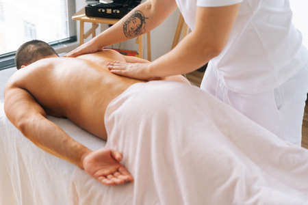 High-angle view of male masseur making gentle stroking movements with strong tattooed hands along spine toward head. Therapist massaging back of muscular sportsman lying on massage table, closeup.の写真素材