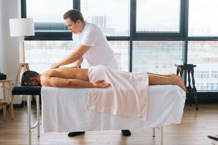 Side wide shot of professional male masseur with strong tattooed hands massaging back and shoulder of muscular sports man lying on stomach at massage table. Concept of healthy lifestyle and restの写真素材