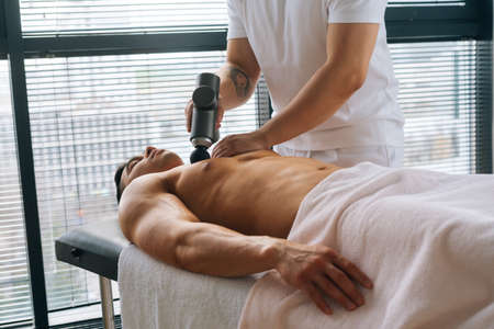 Medium shot of unrecognizable professional male masseur receiving chest muscles using massage gun percussion tool of muscular athlete man, on spa treatment lying on back in massage table.の写真素材