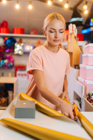 Vertical shot of young woman cutting with scissors wrapping golden paper to package festive cardboard box with Christmas gifts, at counter of holiday store, blurred background of festive packages.の写真素材