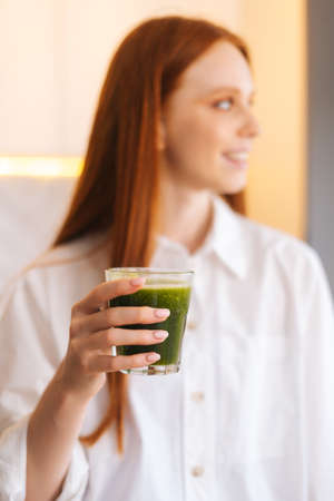 Vertical blurred portrait of cheerful attractive young woman holding glass with green vegetable detox smoothie cocktail from blender in kitchen. Concept of healthy eating and lifestyle.の写真素材