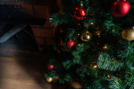 High-angle view of beautiful Christmas tree decorated with festive balls near fireplace at home. Close-up of holiday New Year tree with colorful baubles and illuminated garland in dark living room.の写真素材