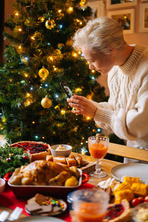Vertical shot of attractive young woman taking photo or making video of beautiful Christmas dinner table on smartphone, on blurred background of decorating xmas tree and celebration bokeh lights.の写真素材
