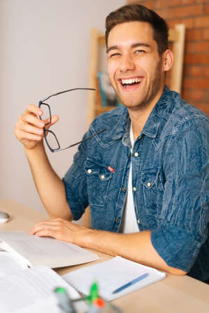 Vertical shot of cheerful laughing young man reading paper book holding end of glasses near mouth sitting at desk in home office, looking at camera. Handsome student male studying alone with textbook.の写真素材