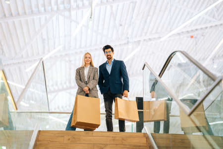 Low-angle view of happy beautiful young couple holding shopping paper bags with purchases and looking at camera, standing on top of stairs in mall. Concept of holiday sales, retail, consumerism.の写真素材