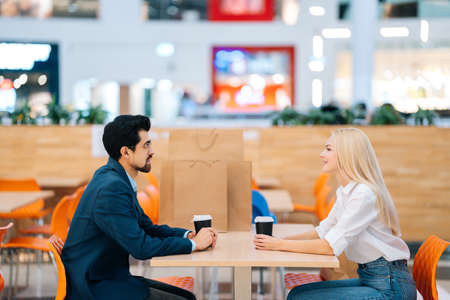 Handsome bearded man and attractive blonde woman having date sitting at table looking at each other in mall. Beautiful young couple drinking coffee and talking in cafe after shopping in store.の写真素材