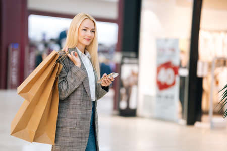Smiling attractive blonde young woman in stylish coat holding mobile phone and shopping paper bags...
