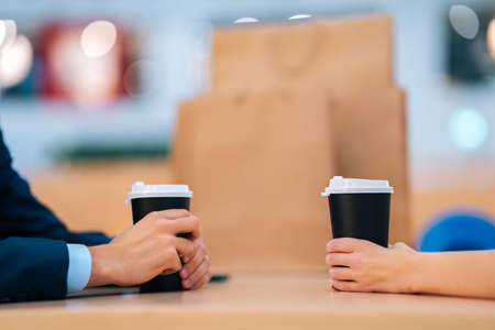 Close-up side view of unrecognizable young man and woman holding cup with coffee sitting at table opposite each other. Young couple drinking coffee and talking in cafe after shopping in store.の写真素材