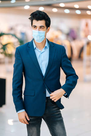 Vertical portrait of confident young man in stylish suit wearing protection face mask looking away in hall of mall centre, blurred background. Front view of handsome Caucasian male applying mask.の写真素材