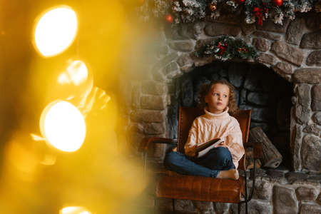 Dreaming pensive little blonde curly child girl writing letter to Santa Claus sitting on chair by fireplace at home with festive interior. Pretty Caucasian kid writes letter with wishes for gift.の写真素材