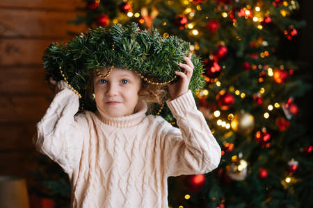 Portrait of adorable little blonde curly child girl holding Christmas standing in dark cozy room with festive interior, on background of decorated Christmas tree with bokeh lights.の写真素材
