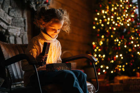 Charming smiling little blonde child girl opening magical Christmas box with gift sitting on rocking chair in xmas morning in dark living room, on background of decorated tree with bokeh lights.の写真素材