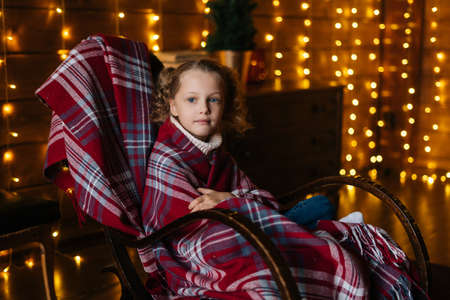 Pretty little blonde curly child girl wrapped in plaid looking at camera sitting in rocking chair at cosy dark room with festive interior, background of wall decorated Christmas bokeh lights.の写真素材
