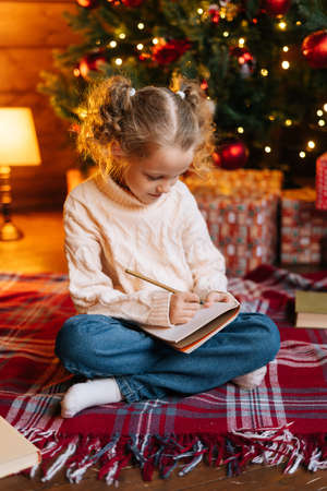 Vertical portrait of focused little blonde curly child girl writing letter to Santa Claus sitting on floor on background of Christmas tree and gift boxes, living room with festive red bright interior.の写真素材