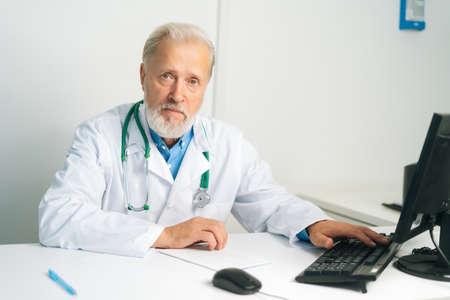 Portrait of serious mature adult male doctor in white coat with stethoscope working on desktop computer sitting at desk in medical office, looking at camera. Confident senior physician at workplace.の写真素材