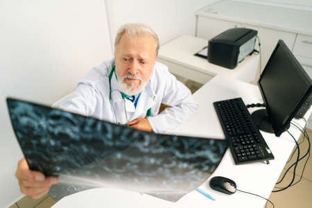 High-angle view of mature adult male doctor examining MRI spine image of patient sitting at desk with computer in medical office room, thinking about diagnosis, expressing concern of illness.の写真素材