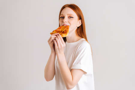 Studio portrait of cheerful happy young woman with appetite to eat delicious pizza looking at camera standing on white isolated background. Pretty redhead female eating tasty meal.の写真素材