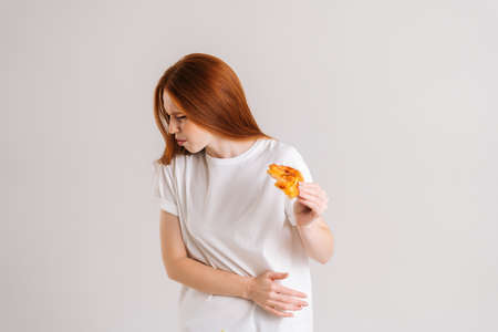 Studio portrait of sick young woman closed eyes feeling pain in stomach after eating pizza, suffering abdomen ache, standing on white isolated background, holding pizza slice.の写真素材