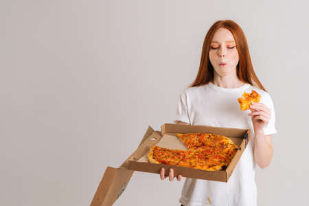 Studio portrait of happy young woman closed eyes with appetite to eat delicious pizza holding box in hands standing on white isolated background. Pretty redhead female eating tasty meal.の写真素材