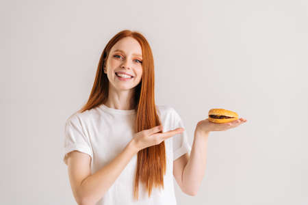 Studio portrait of cheerful pretty young woman pointing to tasty burger and looking at camera standing on white isolated background. Happy cute redhead female eating bad hamburger.の写真素材