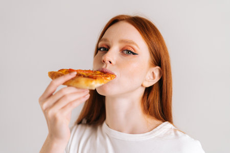 Close-up face of happy attractive young woman with appetite eating delicious pizza looking at camera standing on white isolated background. Pretty redhead female eating tasty meal.の写真素材
