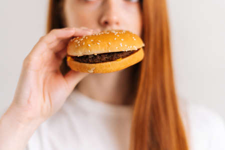 Close-up cropped shot face of unrecognizable young woman pleasing bite of appetizing delicious burger on white isolated background in studio. Closeup front view of female eating tasty hamburger.の写真素材
