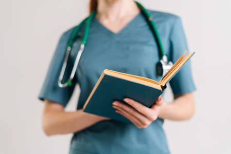 Close-up cropped shot of unrecognizable female doctor intern in uniform with stethoscope reading medical book, standing on white isolated background in studio.の写真素材
