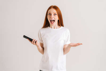 Studio portrait of amazed young woman open mouth reading good online message using mobile phone on white isolated background. Happy redhead lady holding smartphone in hands and looking at camera.の写真素材