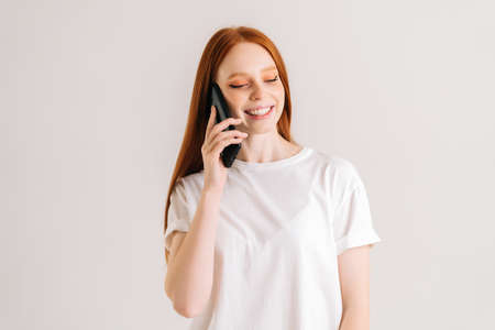 Studio portrait of cheerful redhead young woman having pleasant mobile conversation using smartphone...